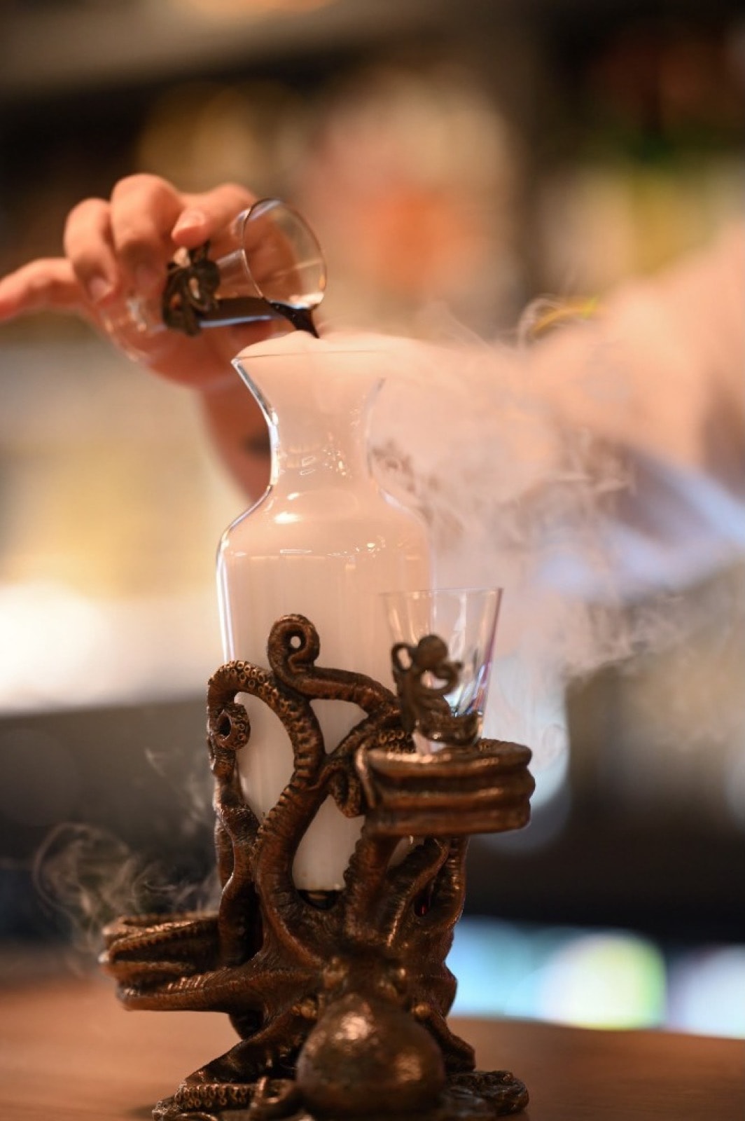 Un bartender vierte un licor oscuro en una jarra de cristal de la que rebosa vapor de hielo seco, sobre un soporte con forma de pulpo