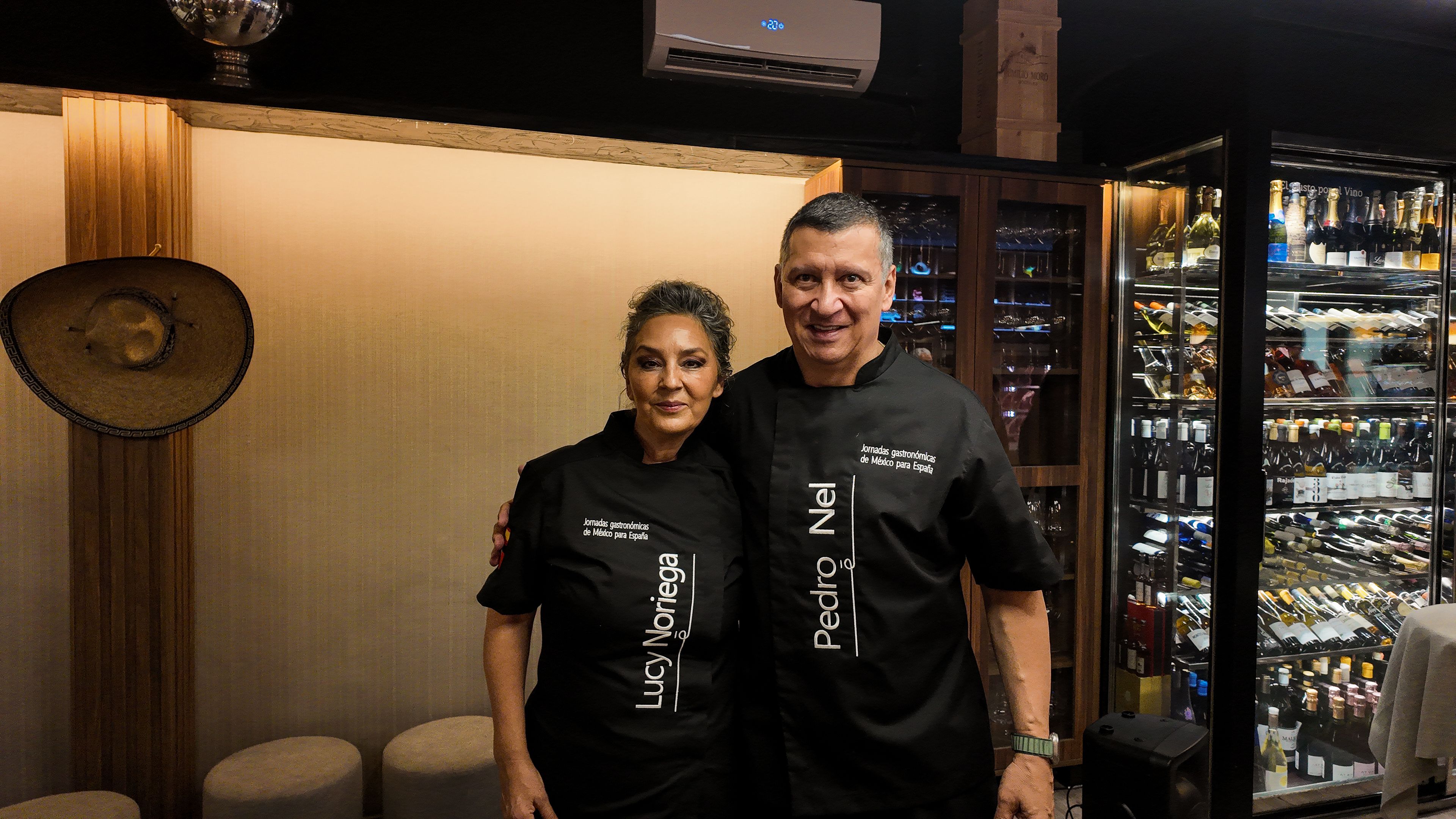 Lucy Noriega y Pedro Nel, abrazados, con las chaquetillas negras de la jornada gastronómica de México con sus nombres bordados, junto a una vinoteca